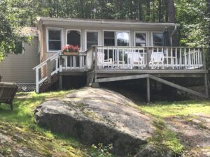 Lakeside view of cottage two showcasing a cottage with many windows, a deck and large rock in the yard