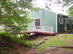 Front view of cottage five showcasing a green cottage with a white deck surrounded by trees